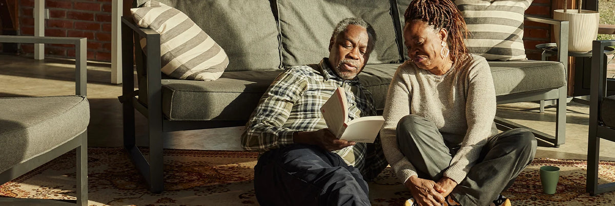 An older man and an older woman sit on sofa and reading a book together. 