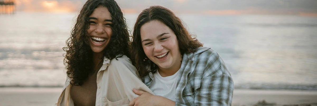 Two young women smiling and laughing together on a beach at sunset.